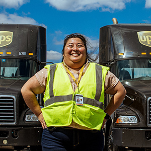 Instructora de seguridad de UPS sonriendo y vistiendo un chaleco de seguridad reflectante de pie delante de dos tractocamiones.