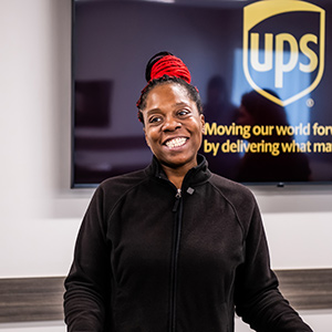 Lächelnde UPS Teilzeitmitarbeiterin vor einer Plakette mit dem UPS Logo.