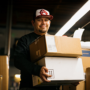 Lächelnder UPS Teilzeitmitarbeiter mit Baseballmütze beim Tragen von  Kartons.