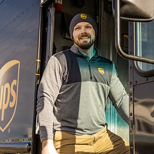 Smiling UPS employee in a branded beanie and leaning out of his delivery truck.