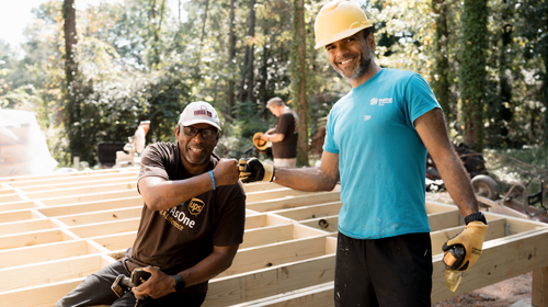 茶色の制服と青い手袋を着用した人が、屋外の工業環境で大型トラックに給油しています。作業員は燃料ホースを取り扱い、トラックのタンクに接続しています。背景にはトレーラーと空があり、作業環境を示しています。この画像は、安全と輸送業務を強調しています。