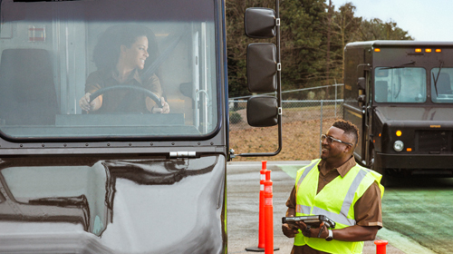 配達トラックが屋外の検問所で停車し、そこで視認性の高いベストを着用した作業員がデジタルタブレットで検査を行っています。