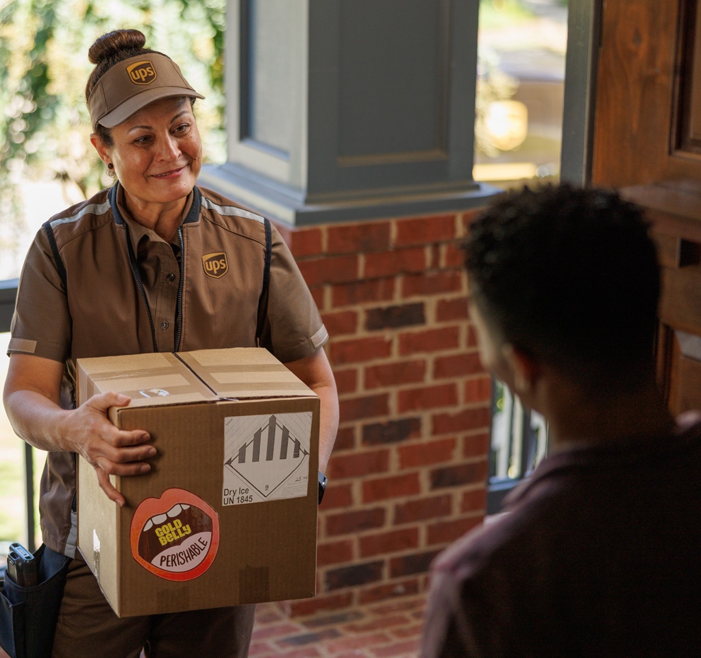 UPS driver stands on a brick porch, holding a large cardboard box labeled 'FRAGILE' 