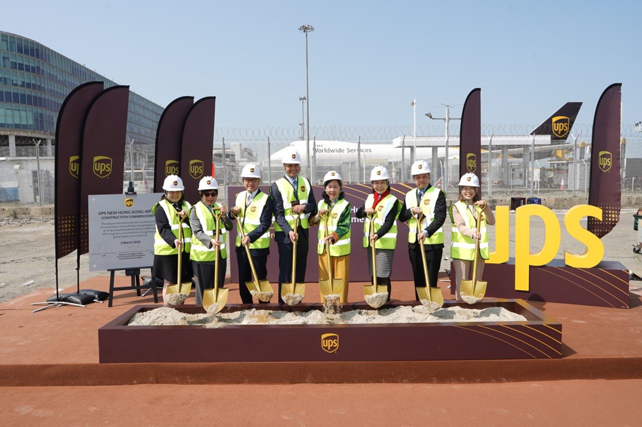 A group of adults in safety vests and hard hats pose with shovels at a UPS groundbreaking ceremony. They stand on a raised platform filled with sand at an outdoor construction site. Multiple UPS flags and a large yellow UPS sign are prominently displayed behind them. An airport or industrial facility with fencing and an airplane is visible in the background under clear daylight.