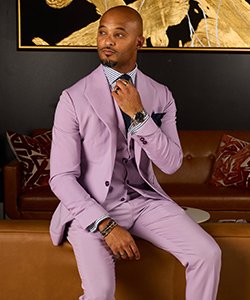 A portrait of Omar Kinnebrew wearing a lavender suit, seated on a leather couch