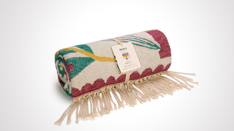 A rolled woven blanket featuring vibrant patterns in red, green, and yellow tones. The blanket includes fringe detailing and is displayed against a clean white background. A visible tag on the blanket highlights the design and branding.
