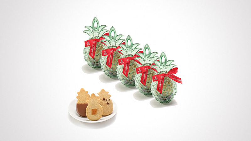 A set of five decorative pineapple-shaped glass jars adorned with red ribbons, arranged in a row. In the foreground, a plate holds pineapple-shaped cookies, including chocolate-filled ones. The image features a clean white background, emphasizing the festive and tropical theme.