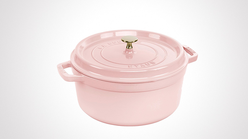 A pastel pink cast iron Dutch oven with a matching lid and gold-tone knob. The product is displayed against a clean white background, highlighting its elegant design and sturdy handles. The word 'STAUB' is embossed on the lid and handles, adding a touch of branding detail.