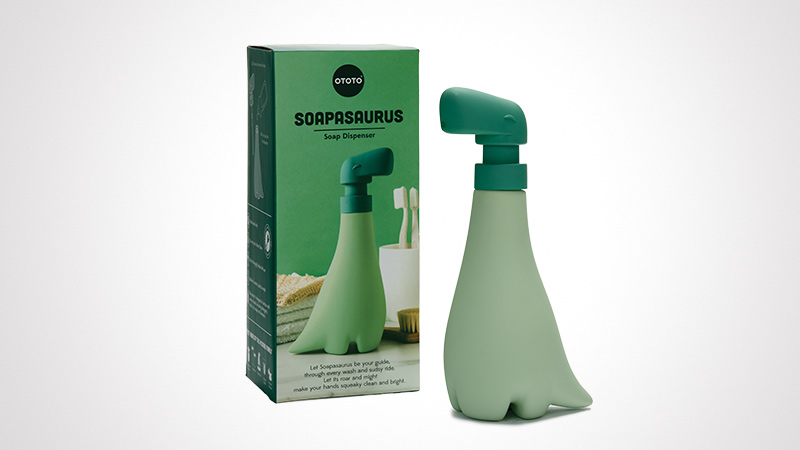 A dinosaur-shaped soap dispenser by Ototo, featuring a playful green design. The product is displayed alongside its packaging, which includes visible branding and text. Ideal for adding a fun touch to bathroom or kitchen settings.
