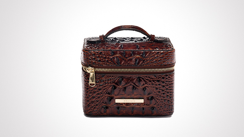 A compact vanity case featuring a rich brown textured design resembling crocodile skin. The case is equipped with a sturdy handle and a gold zipper for secure closure. A metallic plate with visible text 'BRAHMIN' is affixed on the front. The clean white background highlights the luxurious and elegant design.