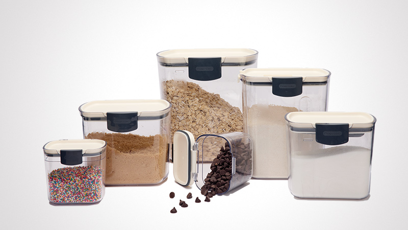 A set of six transparent storage containers filled with various ingredients, including sprinkles, brown sugar, oats, flour, and chocolate chips. The containers feature black lids and are displayed against a plain white background. One container is tipped over, spilling chocolate chips onto the surface.