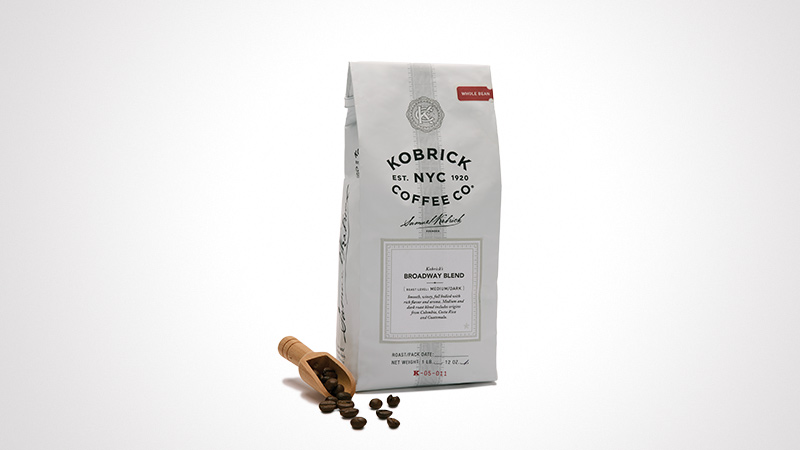 A white coffee bag labeled 'Kobrick Coffee Co.' featuring the Broadway Blend. The packaging includes visible text and design elements, with coffee beans and a wooden scoop placed nearby. The numeric value '12 oz' is clearly displayed on the bag.
