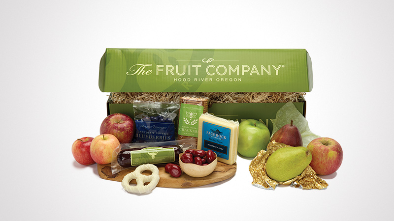 A green gift box from The Fruit Company, Hood River Oregon, containing fresh fruits, cheese, and snacks. The assortment includes apples, pears, cherries, pretzels, and packaged cheese. The box is styled with natural straw filling and features visible branding text.