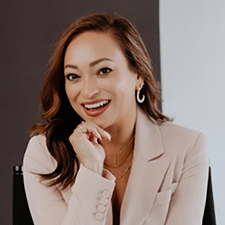 A woman is seated and dressed in a tailored beige blazer, posing thoughtfully with her hand near her face. She wears layered gold necklaces and hoop earrings that add a polished, modern touch. The background is split into dark and light panels, creating a clean, studio-like setting. The overall scene suggests a professional, fashion-forward portrait with a warm, neutral color palette.