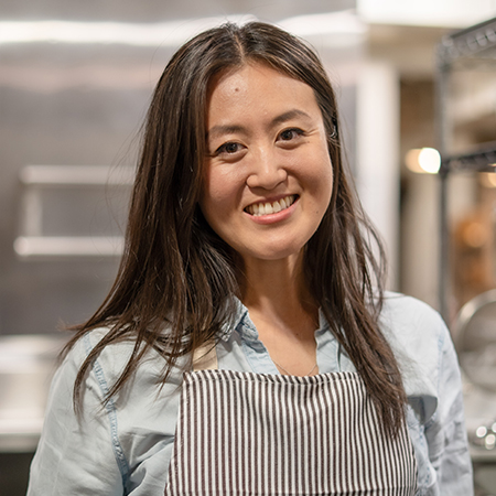 A woman stands in a professional kitchen wearing a striped apron over a light-colored shirt. Her face is obscured, drawing attention to her attire and the culinary setting. Stainless steel equipment and shelves in the background suggest a commercial or restaurant environment. The overall scene conveys a busy, work-focused atmosphere.