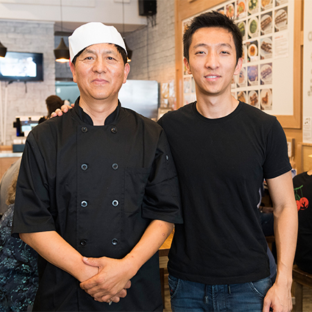 Two adults stand side by side inside a casual restaurant. One person wears a black chef coat and white cap, suggesting they work in the kitchen. The other person is dressed in a plain black t-shirt and jeans, appearing as a guest or friend. The background shows a bright dining area with wall menus, framed food photos, and other patrons seated at tables.
