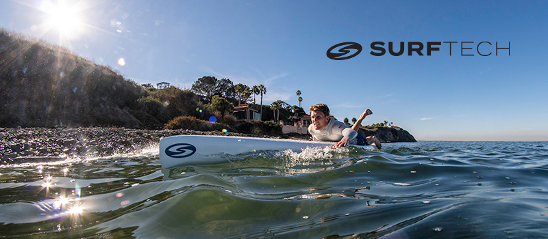 A man is seen paddling on a Surftech board in calm coastal waters. The setting features a sunny day with reflections on the water and a scenic shoreline with houses and palm trees in the background. The Surftech logo is clearly visible on the board and in the overlay text.