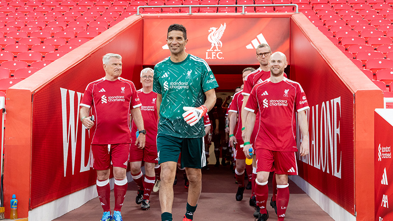A group of soccer players walks out from a stadium tunnel toward the pitch. The central figure wears a green goalkeeper kit while others wear matching red uniforms. The tunnel walls and seating area are bright red, with large white text and symbols visible in the background. The scene suggests a professional or exhibition match in a large arena. The overall atmosphere is energetic and anticipatory as the team enters the field.