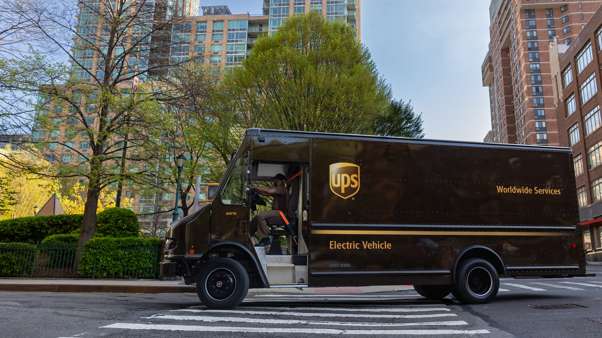 Photo of UPS driver in UPS package car driving down the road with trees and buildings in the background