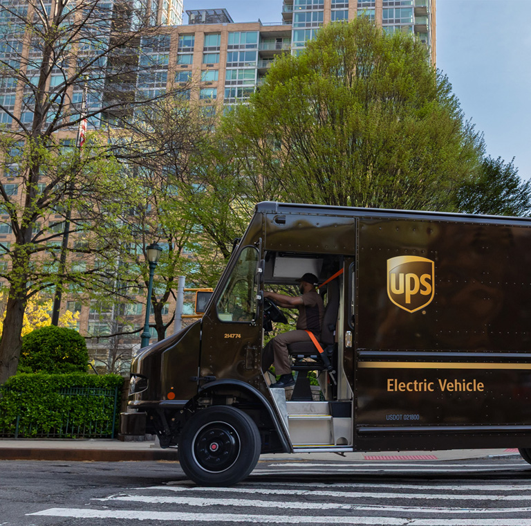 Photo of UPS driver in UPS package car driving down the road with trees and buildings in the background