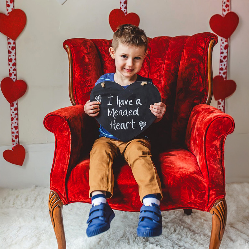 A young child sits on a vibrant red armchair, holding a heart-shaped sign that reads 'I have a mended heart.' The setting features a festive backdrop with red hearts and decorative accents. The child is dressed casually in brown pants and blue shoes, creating a warm and cheerful atmosphere.