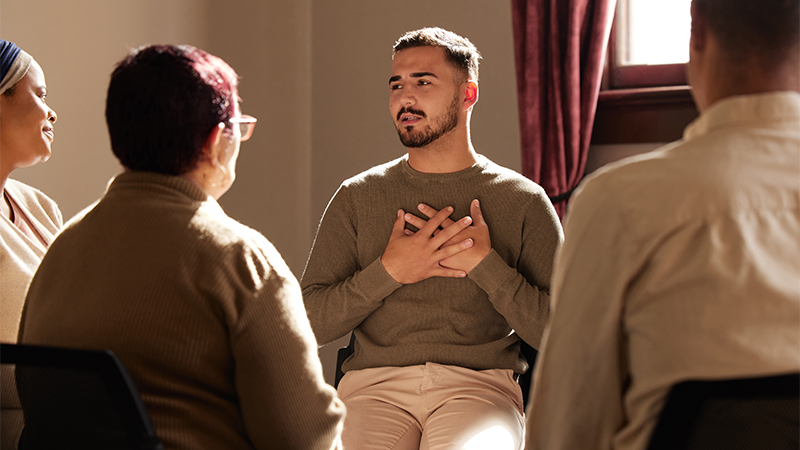 A small group of adults sit in a circle during a support group meeting. A man in the center holds his hands over his chest as he speaks, suggesting an emotional or heartfelt moment. Warm sunlight enters through a nearby window, creating a calm and intimate atmosphere. The participants appear engaged and attentive to the speaker.