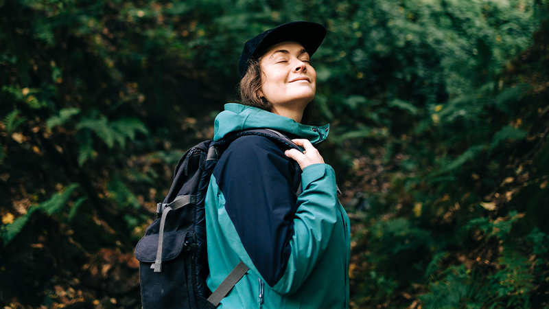 A person wearing a teal and black jacket carries a backpack while walking through a dense forest. The subject is viewed from the side with their face obscured, focusing attention on the outdoor gear. Lush green foliage surrounds the hiker, suggesting a nature trail or woodland hike. The overall scene conveys exploration and adventure in a natural setting.