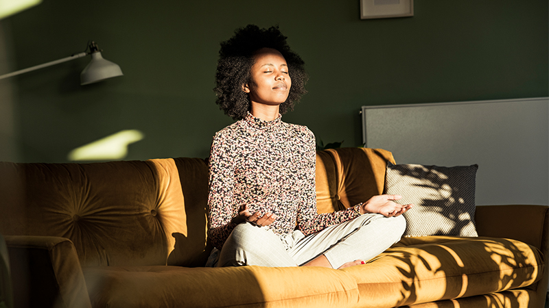 A woman sits cross-legged on a mustard-yellow sofa, practicing meditation. She is indoors in a cozy living room with green walls and soft natural light streaming in. Her hands rest on her knees in a relaxed pose, and she wears a patterned top and light-colored pants. Shadows and warm sunlight create a calm, peaceful atmosphere.