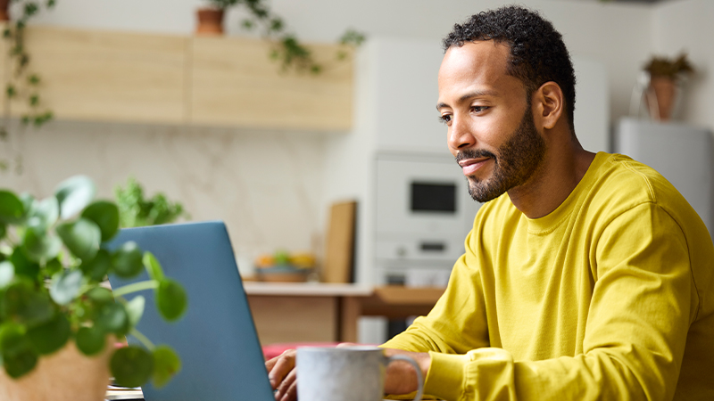 A man in a bright yellow sweatshirt works on a laptop at a table. He is seated in a modern kitchen with light wood cabinets and a built-in oven in the background. Green plants and soft natural light create a relaxed, homey atmosphere. A mug sits on the table, suggesting he may be working or studying from home.