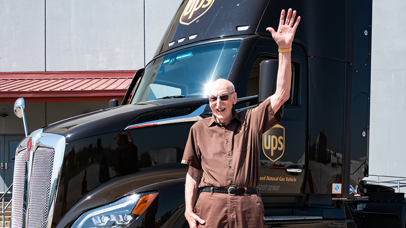 A UPS-branded delivery truck is parked outdoors near a building with a red roof. The truck features the UPS logo prominently on its front and side. Cleve is standing beside the truck, raising one hand in a wave. The truck is labeled as a natural gas vehicle, showcasing eco-friendly transportation.